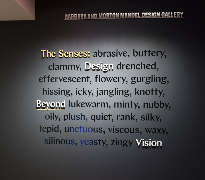 A dark wall features even darker text describing adjectives to describe how we sense our world, the words "The Senses, Design Beyond Vision" are raised from the surface of the dark wall in yellow and white.