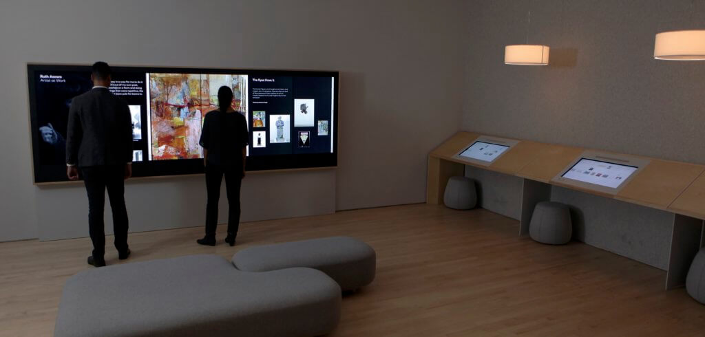 Two people stand in front of a large multi-person, multitouch interface composed of three vertical touchscreens placed side-by-side to form a touchable wall of art in the San Francisco Museum of Modern Art.