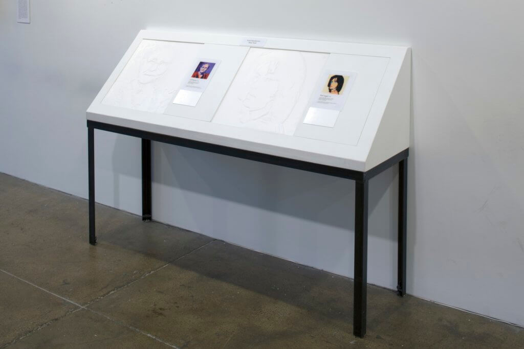 Two tactile images of Andy Warhol paintings are displayed with braille descriptions and a color reproduction of the artwork on a table that is tilted up in a 45 degree angle from the wall to ease the user when they touch the images.