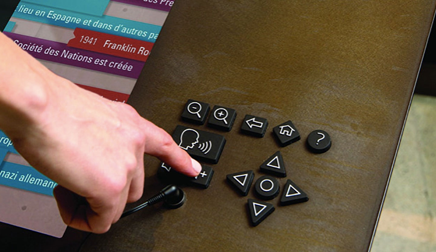 A finger presses the increase volume button on a rubberized keypad with buttons for speak, zoom, back, home, help, and navigation.