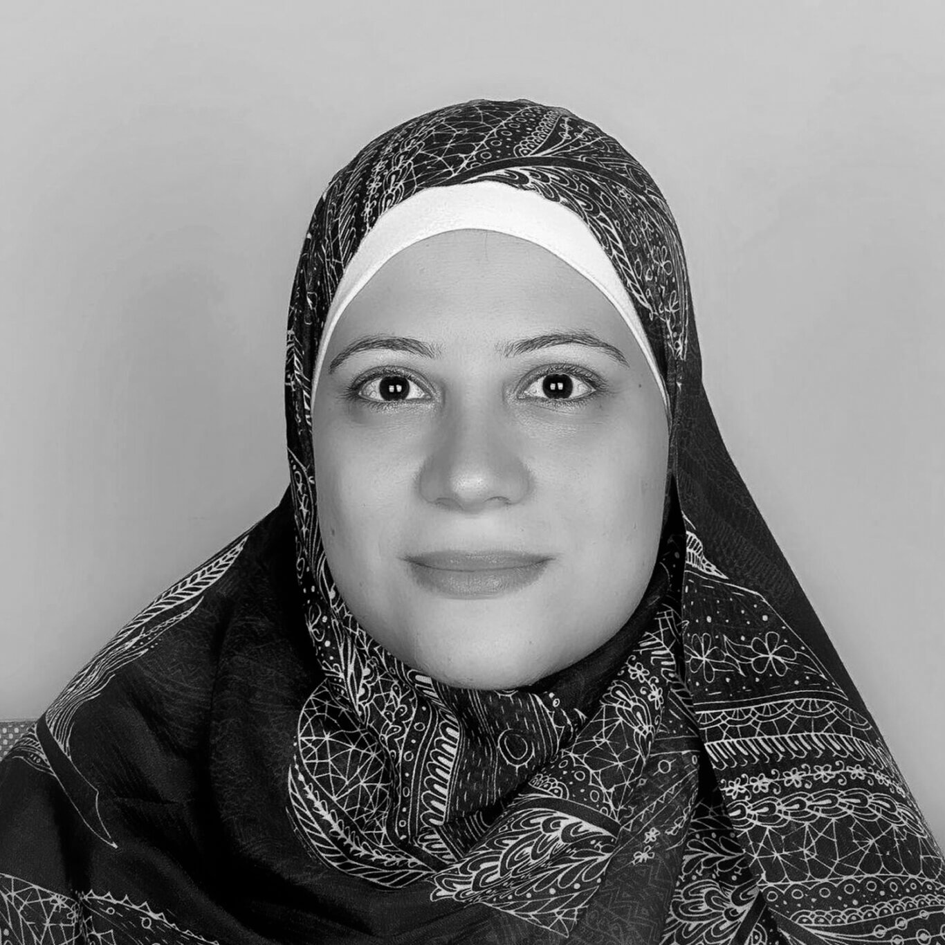 An up close headshot of a woman with light skin tone and a slight smile looks directly at us with dark brown eyes and thin arched eyebrows. She wears a multi toned and patterned hijab. The hijab is wrapped tightly around her head and drapes over her shoulders with soft folds in the fabric. Thin white lines adorn the scarf in geometric and floral patterns. She is in front of a plain light grey wall.
