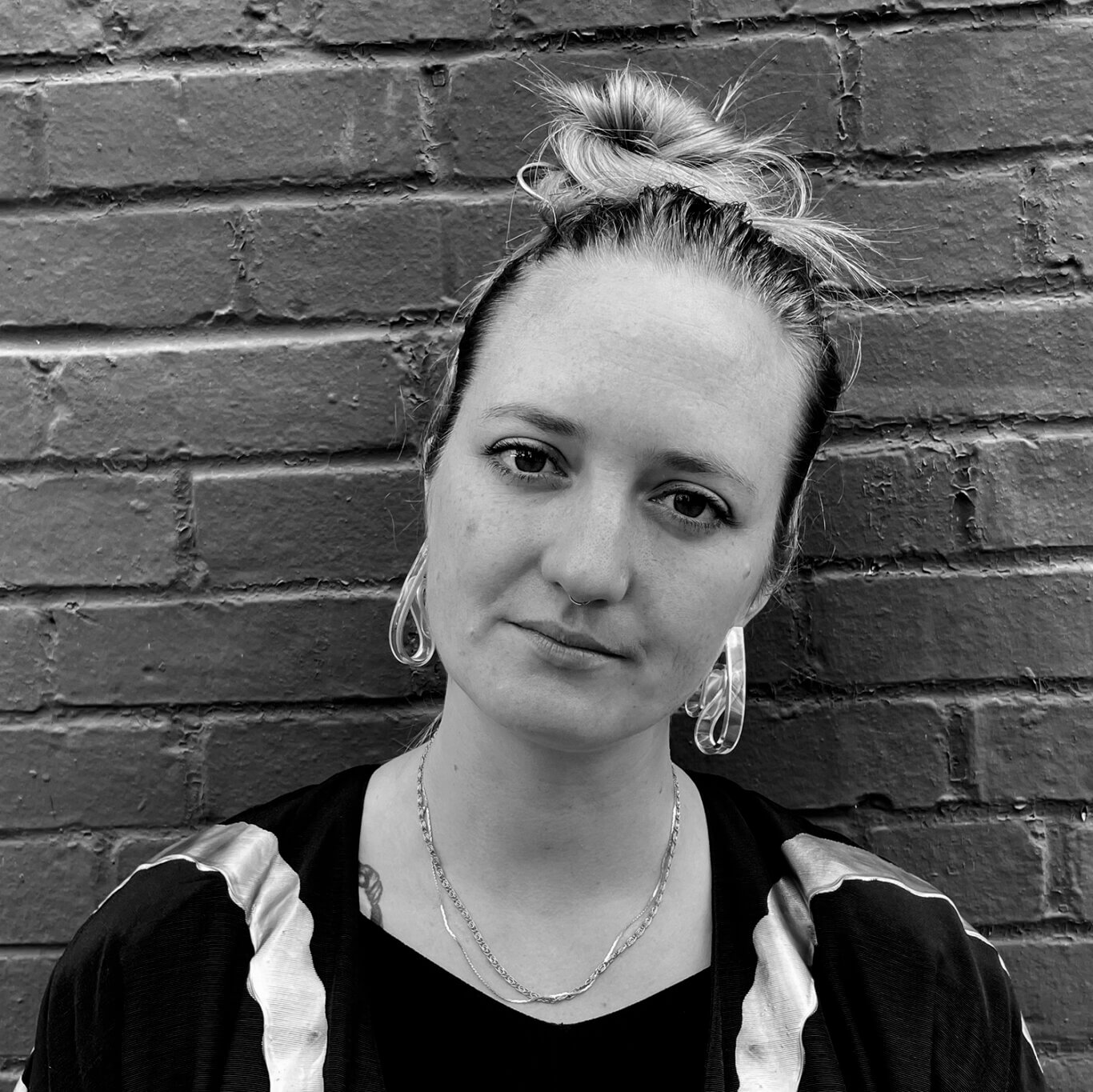 A closely cropped black and white headshot of a White nonbinary femme with light skin smiles slightly at us, their head tilted to our right. Their light hair is in a messy bun with clear abstract earrings, a small septum piercing, and two delicate chain necklaces. The edge of a tattoo of a braid peeks out from under their black jacket near their collar bone on our left. The painted brick wall behind them is slate grey.