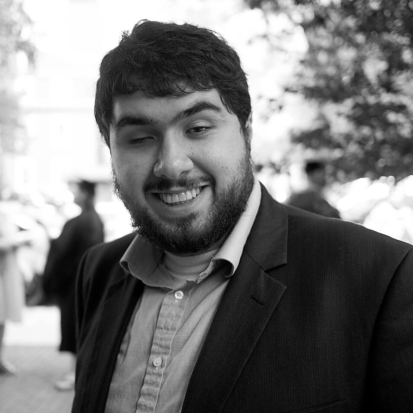 A black and white headshot of a Persian man with light skin and a toothy grin. Shown from the chest up, he has a full beard and thick wavy dark hair. On our left, his right eye is closed, his thick arched eyebrows raised slightly. He wears a charcoal grey sports coat over a light colored button-down shirt. The background is out of focus, with people in graduation gowns and fuzzy foliage.