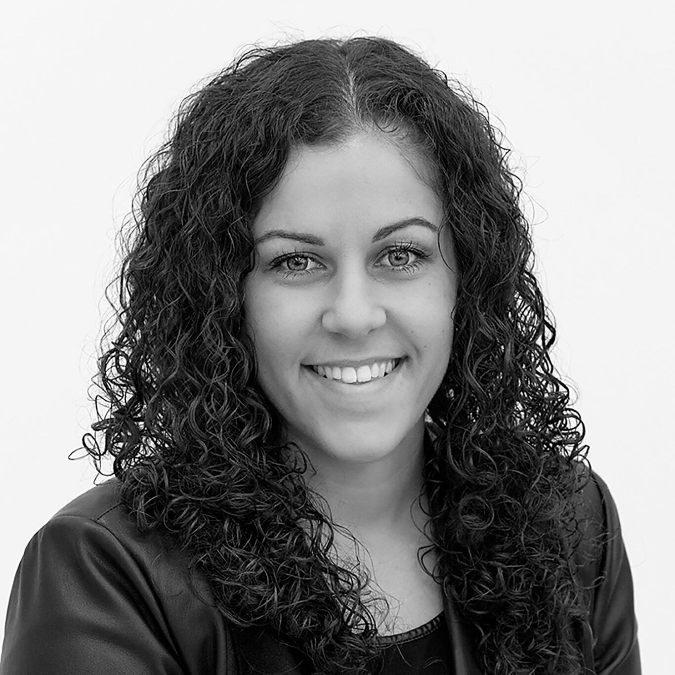 A black and white headshot of a woman with light skin tone, long dark curly hair, long eyelashes, and thin arched eyebrows. Viewed from the chest up, she has a wide smile and wears a black blazer over a black blouse, in front of a white background.